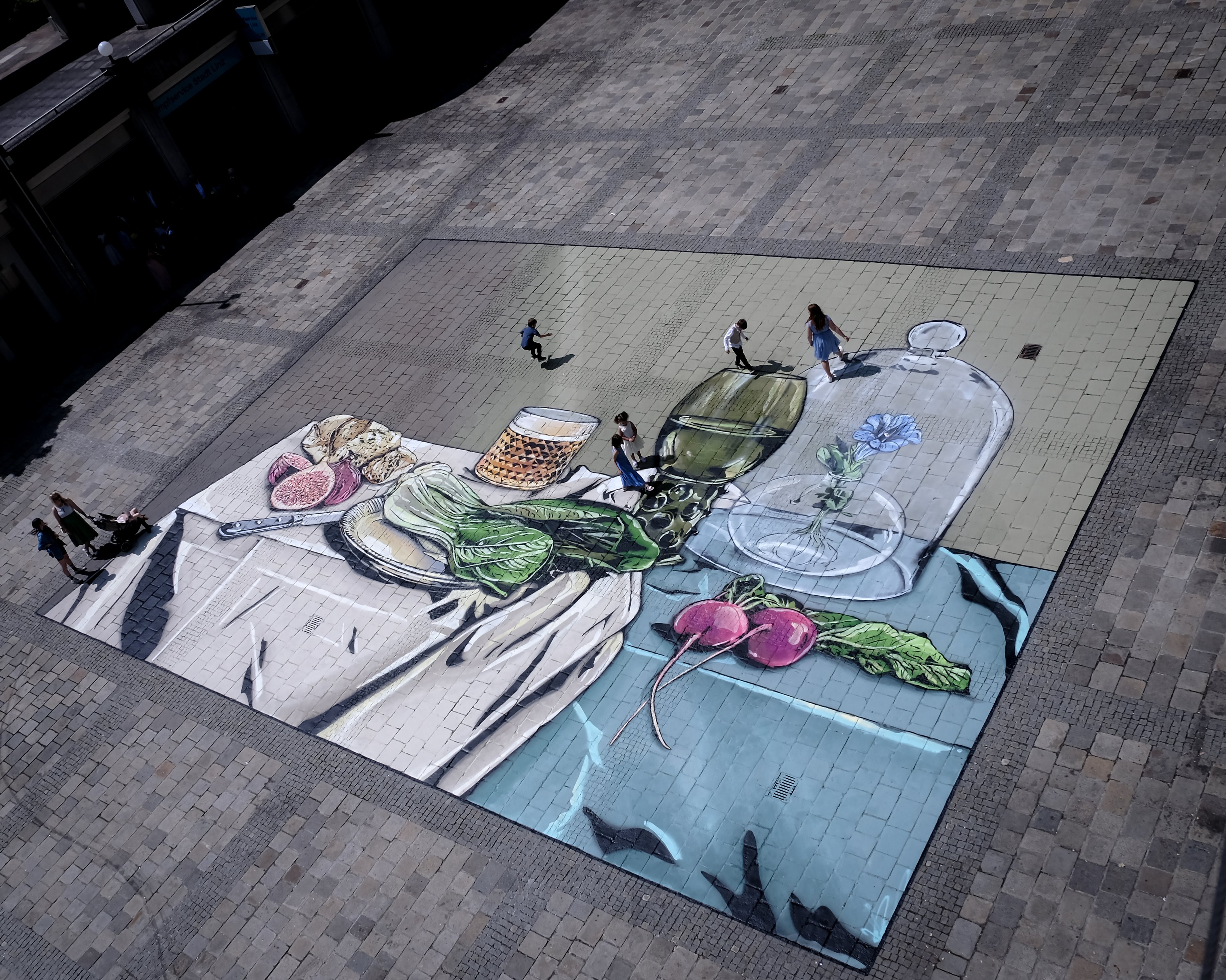 Hier ist ein Foto der Asphalt Art zu sehen, die vor dem Neuen Rathaus in Linz mit Hilfe von Spraydosen aufgebracht wurde. Dabei handelt es sich um eine Neuinterpretation des Gemäldes „Stillleben mit Glaspokal“ von Pieter Claesz. Das Foto wurde von oben aufgenommen und das gesamte Bild ist zu sehen. Im Vergleich zum Originalgemälde fehlen hier der Fisch und die Nüsse, stattdessen sind hier nun Feigen und Salat zu erkennen. Zusätzlich zu sehen ist eine blaue Blume, die in einem Glasgefäß unter einer Käseglocke wächst. Einige Personen bewegen sich gerade über das Gemälde.