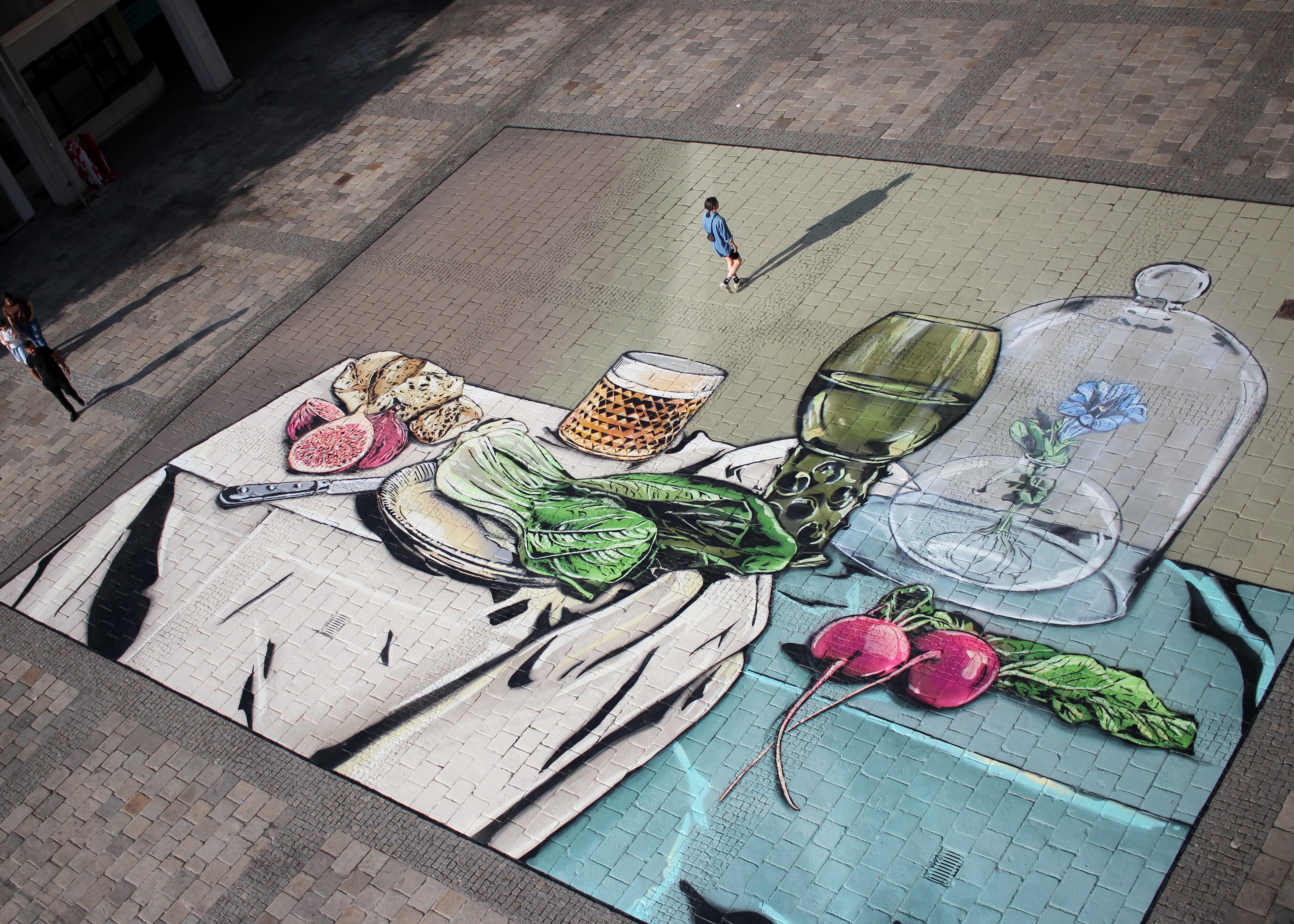 Zu sehen ist ein Foto der Asphalt Art, die vor dem Neuen Rathaus in Linz mit Hilfe von Spraydosen aufgebracht wurde. Dabei handelt es sich um eine Neuinterpretation des Gemäldes „Stillleben mit Glaspokal“ von Pieter Claesz. Eine Person geht gerade über das Asphalt Art, eine andere steht daneben.