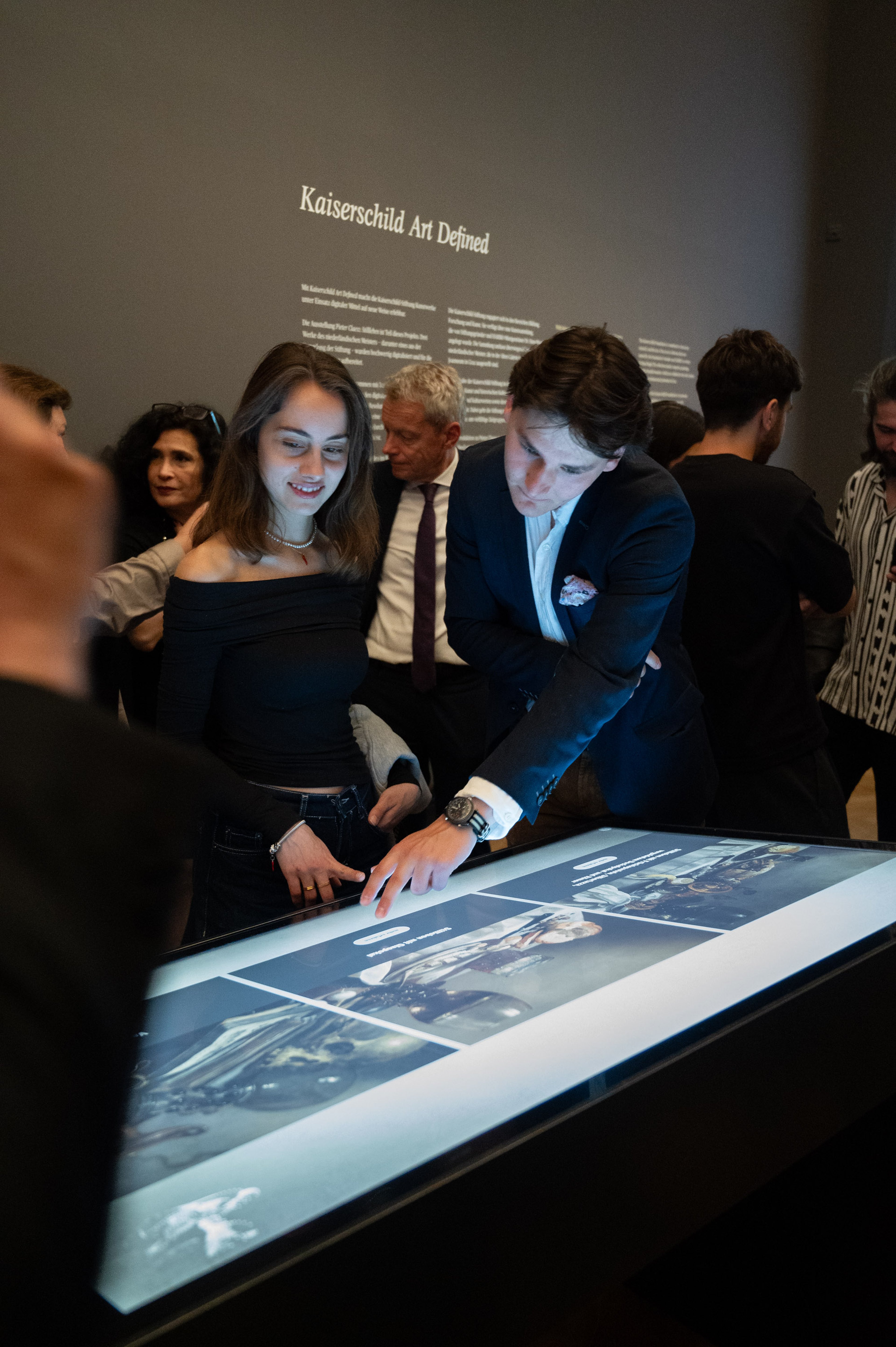 Dieses Foto zeigt zwei Personen, die gerade am Digitalen Tisch in der Ausstellung zugange sind. Im Hintergrund sind einige weitere Personen zu sehen. Die beiden Personen im Fokus sind eine junge Frau und ein junger Mann um die 20 Jahre.