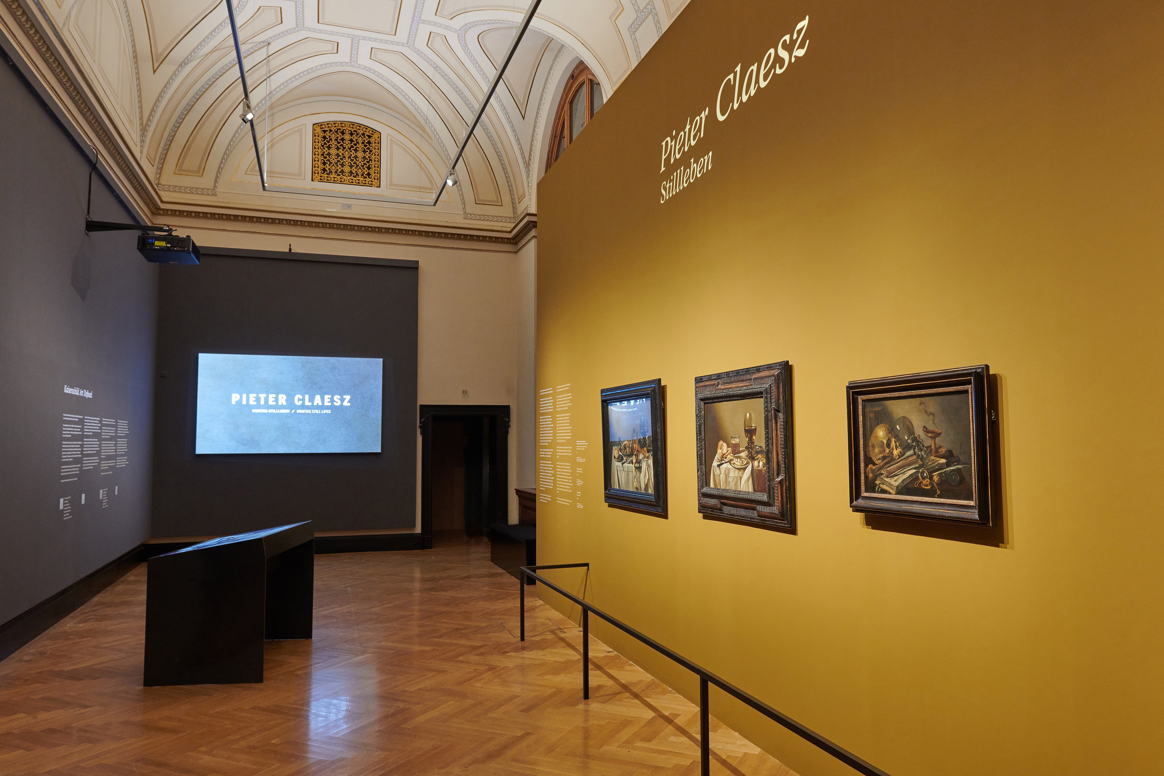 Hier ist ein Ausstellungsraum des Kunsthistorischen Museums zu sehen. An der Wand auf der rechten Seite hängen die drei Gemälde von Pieter Claesz, in der Mitte des Raums steht ein Tisch, an dem die Digitalisate interaktiv erkundet werden können. Ganz im hinteren Bereich des Raums ist eine Projektion des Schriftzugs „Pieter Claesz. Vanitas Stillleben // Vanitas still lifes“ zu sehen.