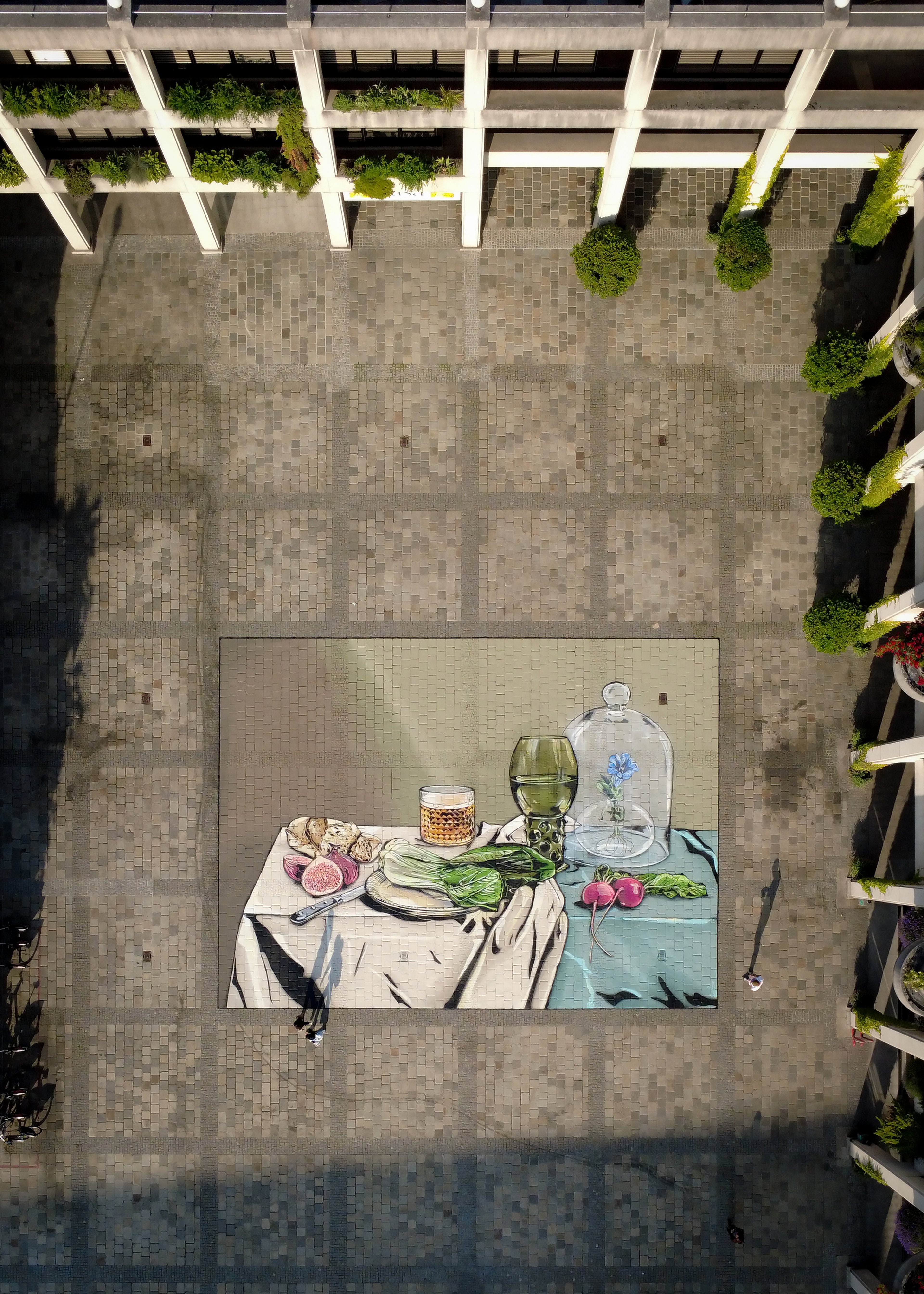 Zu sehen ist ein Foto der Asphalt Art, die vor dem Neuen Rathaus in Linz mit Hilfe von Spraydosen aufgebracht wurde. Dabei handelt es sich um eine Neuinterpretation des Gemäldes „Stillleben mit Glaspokal“ von Pieter Claesz. Das Foto wurde von sehr hoch oben aufgenommen, weshalb auch der übrige Platz sowie Häuserwände zu sehen sind.
